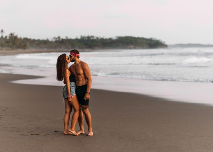 A beautiful couple kissing on the seashore. A couple in love on the coast of Bali. A man and a woman kissing at sunset on the beach. Tanned couple kissing on a sandy beach. Copy space